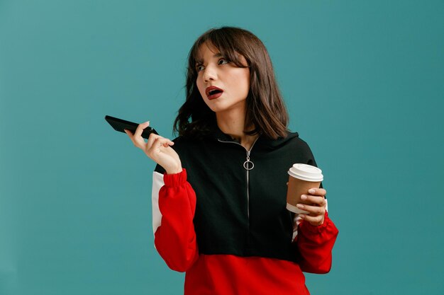 携帯電話と青い背景で隔離の側を見て持ち帰りのコーヒーカップを保持している退屈な若い白人女性