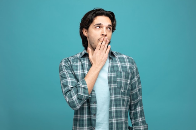 bored young handsome man looking up keeping hand on mouth yawning rolling eyes isolated on blue background