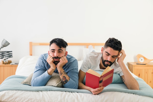 Bored young man lying on bed with his boyfriend reading book