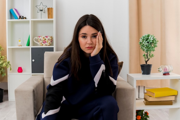 Bored young pretty caucasian woman sitting on armchair in designed living room keeping hand on face and looking