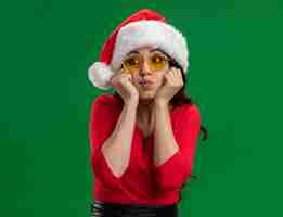 Free photo bored young pretty girl wearing santa hat and glasses keeping hands on chin puffing cheeks looking at side isolated on green background with copy space