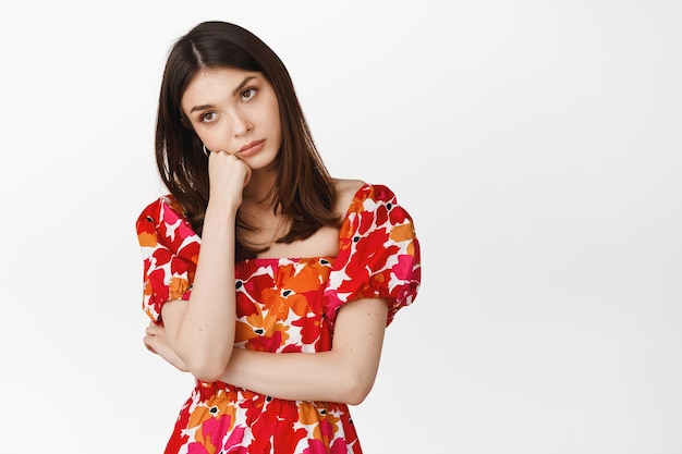 Bored young woman lean head on hand staring bothered or annoyed feeling sad standing in red dress against white background