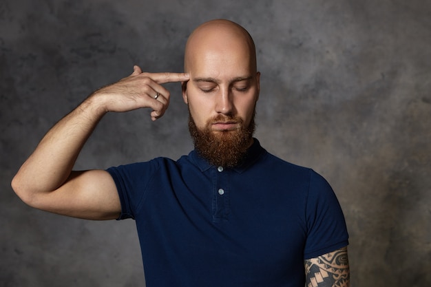 Boredom, fatigue and depression concept. Isolated shot of depressed unhappy Caucasian male with thick beard holding fore finger at his temple and closing eyes as if shooting, feeling stressed