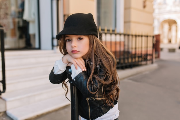 Boring lovely little girl in cute black hat waiting for mother outside in front of beauty saloon.