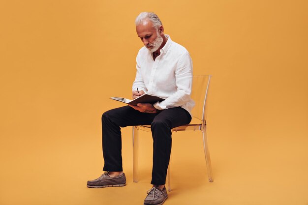 Boss sitting on orange background and makes notes Serious man with gray beard in white shirt is writing something in notebook