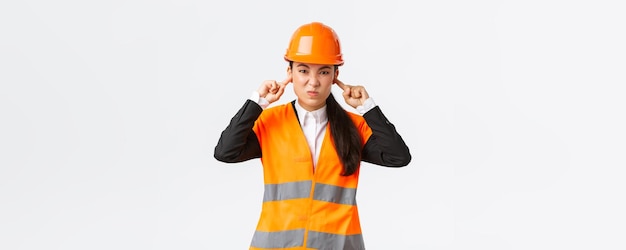 Bothered and displeased asian female chief engineer shut ears and grimacing from awful loud noise at construction area wearing safety helmet complaining on bothering sound white background