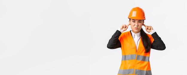 Bothered and displeased asian female chief engineer shut ears and grimacing from awful loud noise at construction area wearing safety helmet complaining on bothering sound white background
