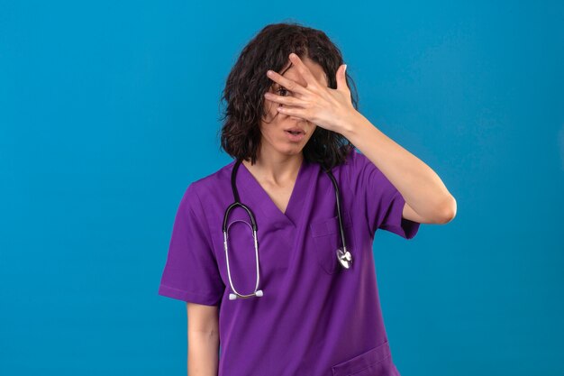 Bothered fed up young nurse wearing uniform and stethoscope coning face with palm looking through palm waiting for surprise standing on isolated blue