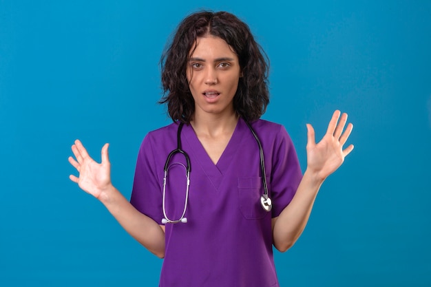 Bothered fed up young nurse wearing uniform and stethoscope standing with hands raised on isolated blue