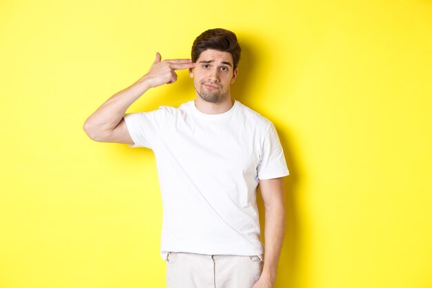 Bothered man shooting himself with finger gun, looking displeased and tired, standing against yellow background.