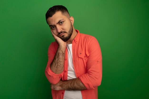 Bothered young bearded man in orange shirt looking  leaning head on palm waiting standing over green wall