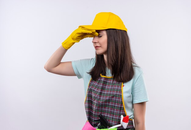 Bothered young beautiful girl wearing apron, cap and rubber gloves closing nose between closed eyes 