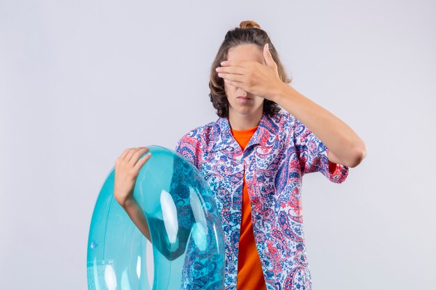 Bothered young handsome guy wearing red sunglasses holding inflatable ring covering face with hand standing 