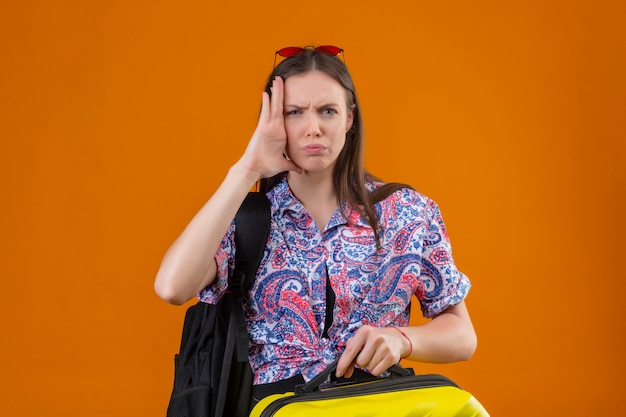 가방을 들고 가방을 들고 서있는 머리에 빨간 선글라스를 착용 한 젊은 여행자 여자가 스트레스를 받고 오렌지에 손으로 머리를 만지고 짜증이납니다.