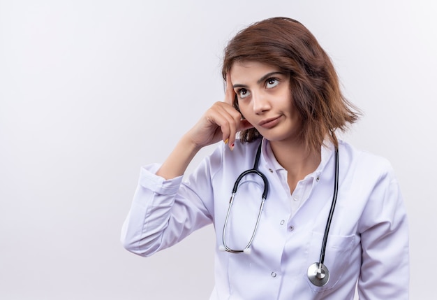 Беспокоит молодая женщина-врач в белом халате со стетоскопом, указывая пальцем на ее висок
