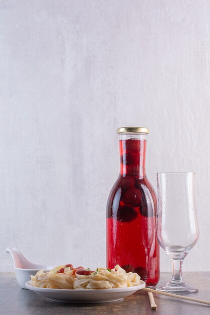 Bottle of red juice with empty glass and pasta on white surface