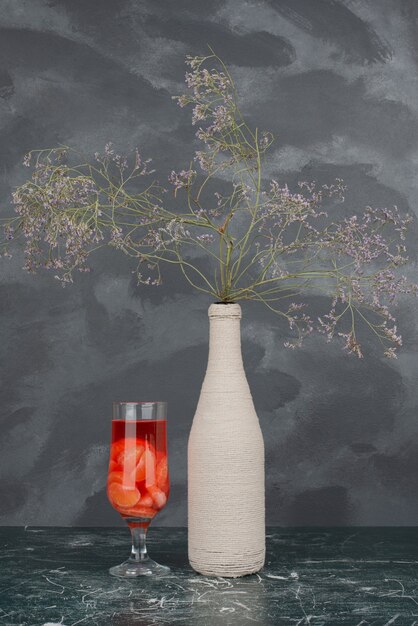 Bottle with withered flowers and glass of juice on marble wall .