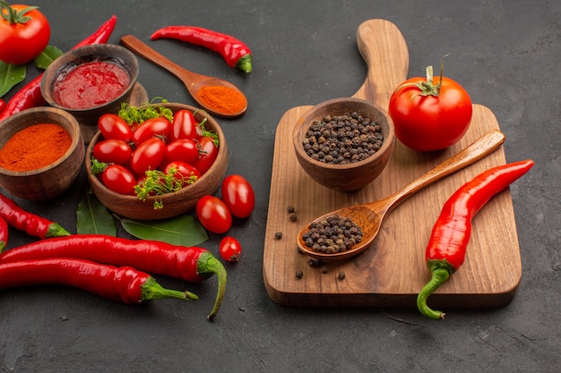 Bottom close view a bowl of cherry tomatoes hot red peppers bay leaves and a bowl of black pepper a wooden spoon a tomato a red pepper on the chopping board on black ground