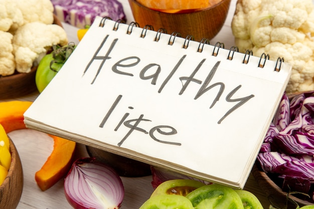 Bottom close view healthy life written on notebook cut green tomatoes cut red cabbage cut pumpkin cauliflower cut bell peppers in bowls on wooden surface free space