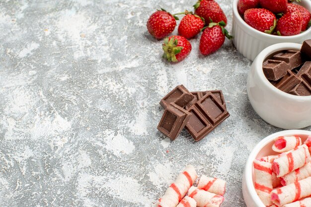 Bottom half view bowls with strawberries chocolates candies and some strawberries chocolates candies at the right side of the grey-white mosaic background