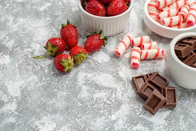Bottom half view bowls with strawberries chocolates candies and some strawberries chocolates candies at the right side of the grey-white table