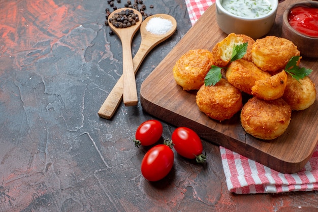 Bottom half view chicken nuggets on wood board with sauces cherry tomatoes wooden spoons black pepper on dark table free space