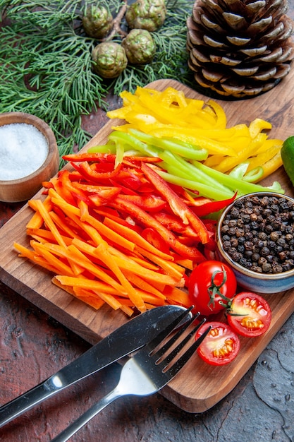 Bottom half view colorful cut peppers black pepper tomatoes on cutting board salt fork and knife pine branches on dark red table