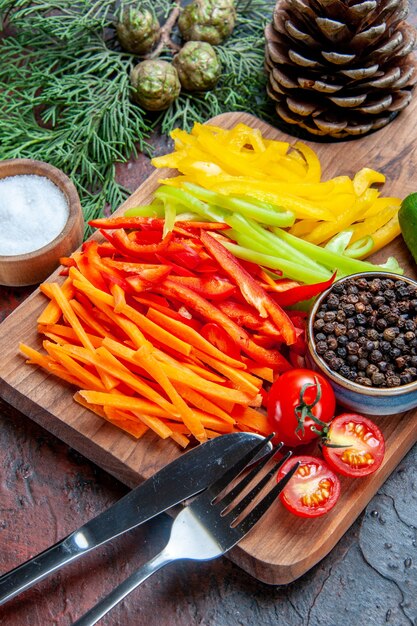 Bottom half view colorful cut peppers black pepper tomatoes on cutting board salt fork and knife pine branches on dark red table