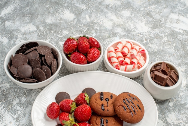 Bottom half view cookies strawberries and round chocolates on the white oval plate surrounded bowls with candies strawberries and chocolates on the background