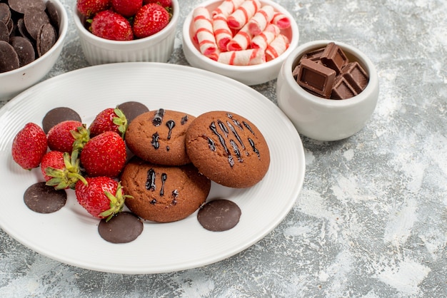 Free photo bottom half view cookies strawberries and round chocolates on the white oval plate surrounded bowls with candies strawberries and chocolates on the grey-white table