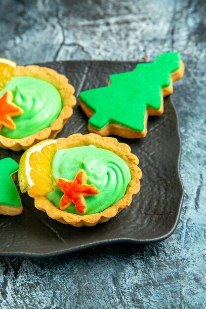 Bottom half view small tarts with green pastry cream xmas tree biscuits on black plate on grey surface