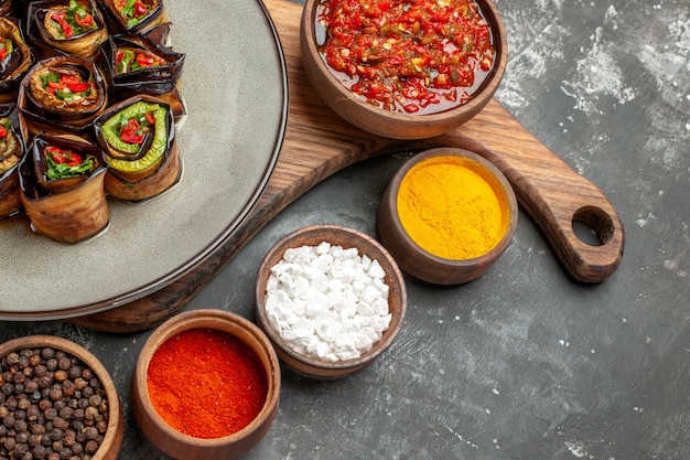 Bottom half view stuffed aubergine rolls in oval plate adjika in bowl on wooden serving board with handle different spices in small bawls on grey background