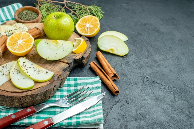 Bottom view apple slices cinnamon sticks and lemon slices apple on wood board pine tree branches a fork and knife on green napkin on black table with free space