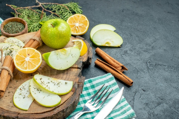 아래쪽 뷰 사과 조각 묶인 계피 스틱과 레몬 조각 사과 나무 판자에 민트를 넣은 소나무 나뭇가지에 포크와 칼을 복사 장소가 있는 검정 테이블에 있는 녹색 냅킨에