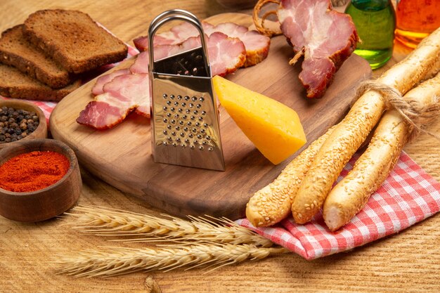 Bottom view becon slices cheese grater on cutting board bread wheat spike red and black pepper in small bowl on brown surface