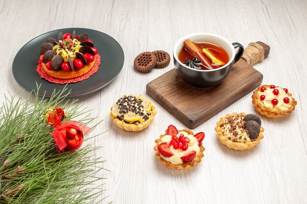 Bottom view berry cake in a grey plate tarts a cup of lemon cinnamon tea on the chopping board cookies and the pine tree leaves with christmas toy on the white wooden background