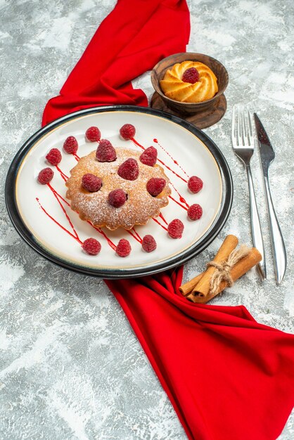 Bottom view berry cake on white oval plate red shawl biscuit fork and dinner knife cinnamon sticks on grey surface