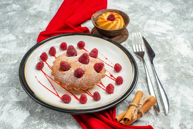 Вид снизу ягодный торт на белой овальной тарелке Красная шаль, вилка для печенья и обеденный нож, палочки корицы на серой поверхности