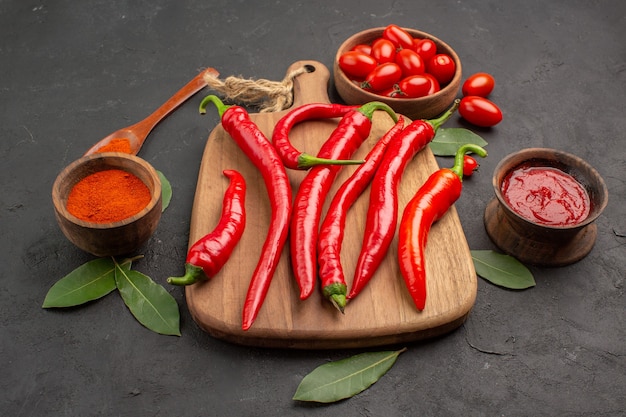 Bottom view a bowl of cherry tomatoes hot red peppers on the chopping board a wooden spoon bay leaves and bowls of ketchup and hot pepper powder on the black table