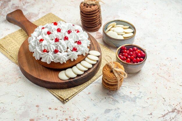 Bottom view cake with white pastry cream on wood board on newspaper berries and white chocolate in bowls cookies tied with rope on light grey surface