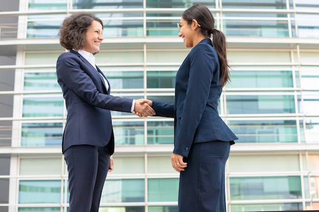 Bottom view of cheerful colleagues shaking hands near building. Young women wearing formal suits meeting outdoor. Business handshake concept