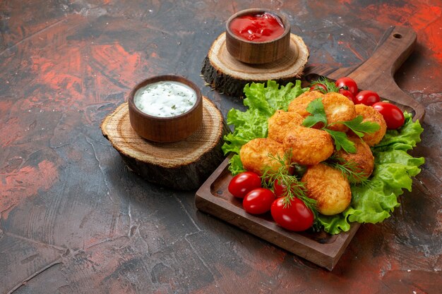 Bottom view chicken nuggets lettuce cherry tomatoes on wood board sauces in bowls on wood boards on dark table