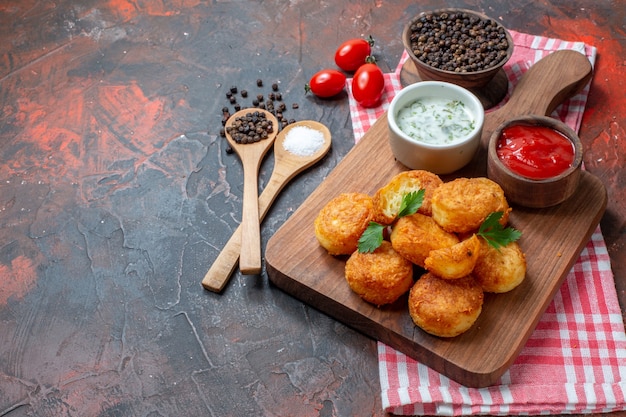 Bottom view chicken nuggets on wood board with sauces cherry tomatoes wooden spoons black pepper in bowl on dark table