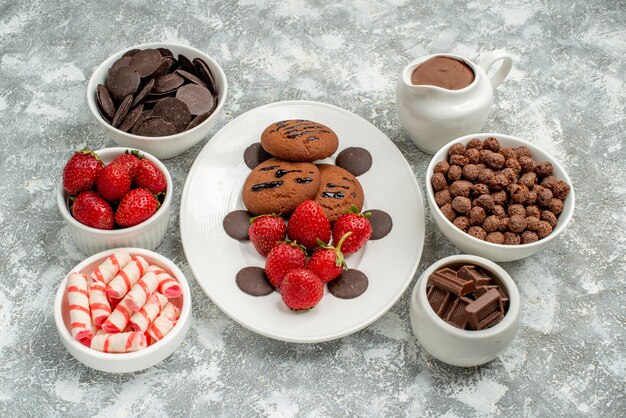 Free photo bottom view chocolate cookies strawberries and round chocolates on the white oval plate and bowls with candies strawberries chocolates cereals and cacao on the grey-white background