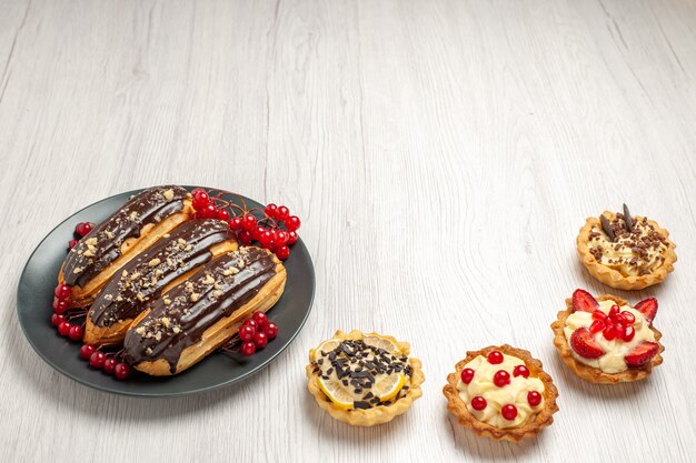 Bottom view chocolate eclairs and currants on the grey plate and four tarts on the white wooden table