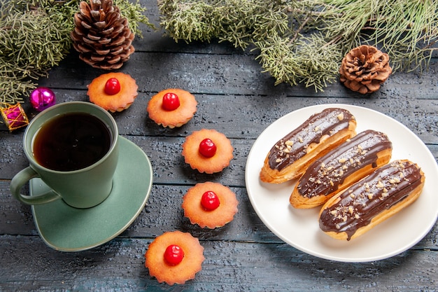 Бесплатное фото Вид снизу шоколадные эклеры на белой овальной тарелке еловые ветки рождественские игрушки кексы и чашка чая на темной деревянной основе