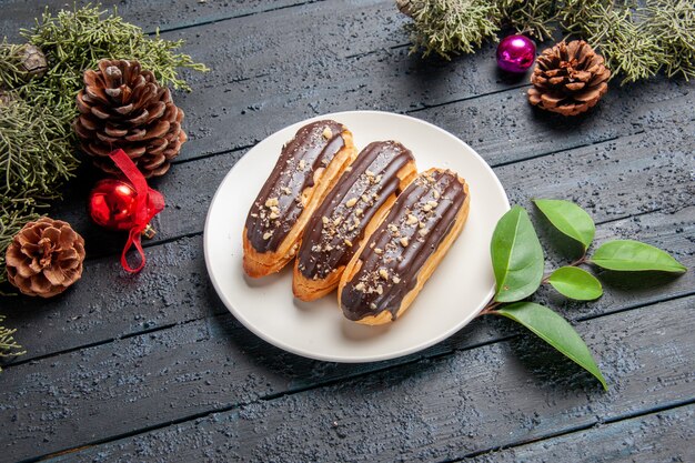 타원형 접시에 밑면 초콜릿 eclairs pinecones 크리스마스 장난감 전나무 나무 어두운 나무 바닥에 나뭇잎