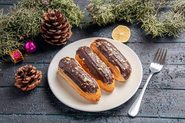 Bottom view chocolate eclairs on white oval plate cones fir-tree leaves christmas toys slice of lemon and a fork on dark wooden table