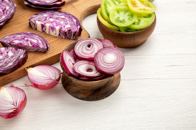 Bottom view chopped onion in bowl cut red cabbage on wood board cut red tomatoes in bowl on white surface