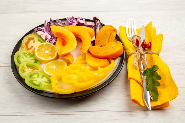 Bottom view chopped vegetables and fruits pumpkin bell peppers persimmon green tomatoes red cabbage on black plate fork and knife on yellow napkin on white surface